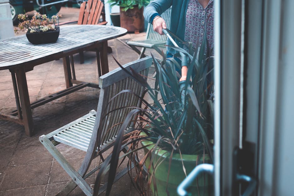 The image depicts a person engaged in garden maintenance on a small outdoor patio area, where a wooden table with potted plants and a matching wooden chair are visible. The individual, partially visib